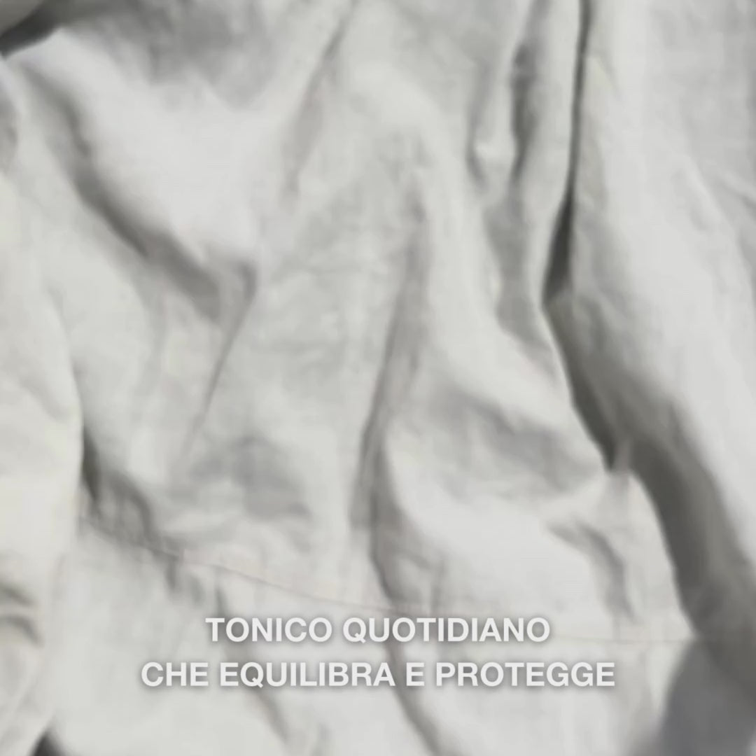Primo piano di una mano che applica alcune gocce del tonico viso trasparente e fluido con Tea Tree Oil su un dischetto di cotone e lo stende delicatamente sulla pelle pulita del viso: il liquido leggero e rinfrescante lenisce, deterge in profondità, idrata e lascia la pelle rigenerata e pronta per la crema.