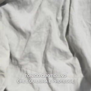 Primo piano di una mano che applica alcune gocce del tonico viso trasparente e fluido con Tea Tree Oil su un dischetto di cotone e lo stende delicatamente sulla pelle pulita del viso: il liquido leggero e rinfrescante lenisce, deterge in profondità, idrata e lascia la pelle rigenerata e pronta per la crema.